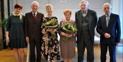 Sala konferencyjna. Trzy pary seniorów stoją obok siebie w rzędzie, kobiety w rękach trzymają bukiety białych kwiatów. 