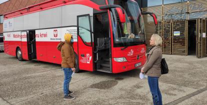  Duży czerwony ambulans stoi przed budynkiem z czerwonej cegły. Jego drzwi są otwarte. Przed wejściem do niego stoją kobieta i mężczyzna. 