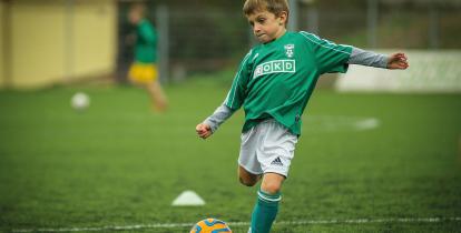 Boisko piłkarskie, na pierwszym planie dziecko w stroju piłkarskim kopiące piłkę nożną.