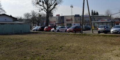 Przestrzeń miejska. Na pierwszym planie fragment działki, w tle parking samochodowy.