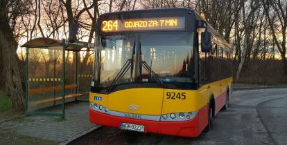 Pomarańczowo-czerwony autobus stoi na przystanku, nad przednią szybą wyświetlany jest numer linii 264 i informacja: odjazd za 7 minut..