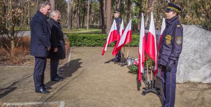 Trzech mężczyzn ubranych na czarno stoi przed pomnikiem katyńskim – dużym kamieniem, na którym wisi granitowa tablica. Pod tablicą leżą kwiaty i palą się znicze. 