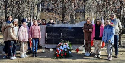 Grupka dzieci oraz dorosłych stoi obok pomnika katyńskiego – dużego kamienia, na którym wisi granitowa tablica. Pod tablicą leżą kwiaty i palą się znicze.