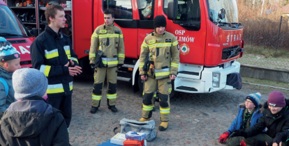 Trzech stojących strażaków na tle wozu strażackiego. Dookoła grupa młodzieży, na ziemi leży niebieska mata, a na niej manekin do ćwiczeń z pierwszej pomocy.