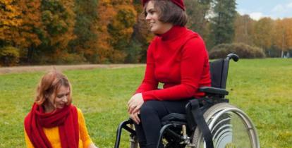 Polana, w tle las jesienią. Na wózku inwalidzkim siedzi kobieta z niepełnosprawnością (uśmiecha się). Towarzyszy jej druga kobieta, która siedzi na kocu rozłożonym na ziemi.