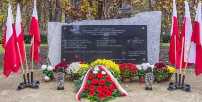 Pośrodku czarna tablica granitowa z tekstem umocowana na prostokątnym kamieniu, po lewej i prawej stronie stoją flagi narodowe, przed tablicą leżą kwiaty i wiązanki oraz palą się znicze