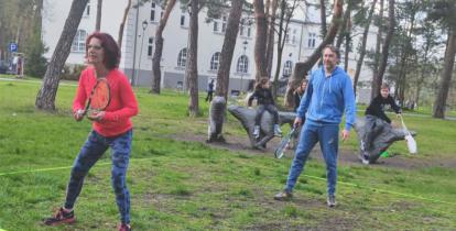 Dwie osoby z rakietkami grają na trawie w crossmintona, w tle dzieci obserwują grę.