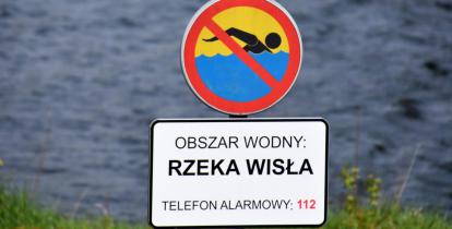 Znak zakazujący kąpieli z tabliczką informacyjną, na której jest tekst: obszar wodny – rzeka Wisła, telefon alarmowy 112. W tle nieostre fale płynącej rzeki.  