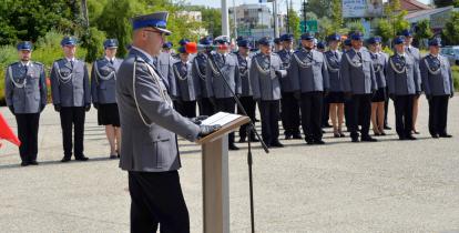 Funkcjonariusze policji w umundurowaniu stoją na baczność w rzędzie. Przed nimi mężczyzna w umundurowaniu przemawia do mikrofonu.