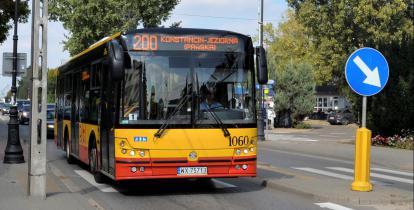 Ulica, po której jedzie czerwono-pomarańczowy autobus komunikacji miejskiej. Nad jego przednią szybą wyświetla się napis: 200 Konstancin-Jeziorna.