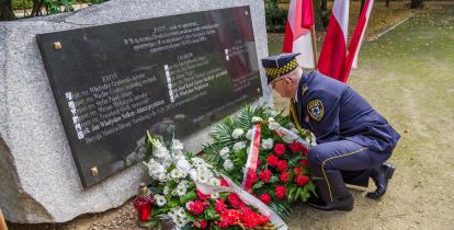  Park. Pomnik ofiar katyńskich. Strażnik miejski składa przed nim wiązankę z białych kwiatów. Obok stoją znicze i trzy biało-czerwone flagi na stojaku. W tle zielone drzewa.