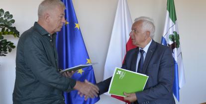 Dwóch mężczyzn przekazuje sobie teczkę na dokumenty. W tle flagi Unii Europejskiej, Polski i Konstancina-Jeziorny.