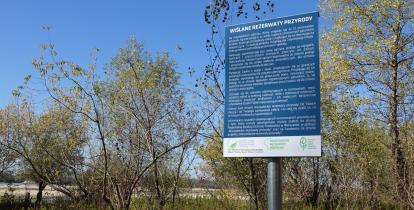 Niebieska tablica a niej białą czcionką podane są liczne zasady obowiązujące na terenie rezerwatu. W tle widać drzewa i rzekę.  