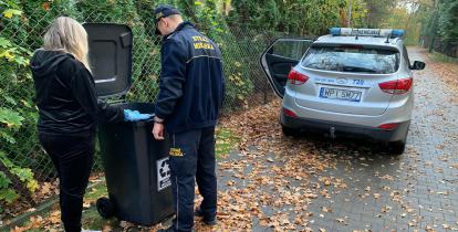 Na zdjęciu kobieta i mężczyna w policyjnym mundurze, zagladają do kosza z odpadami komunalnymi