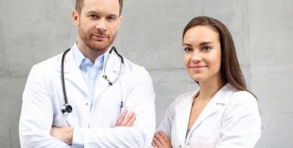 Po lewej stronie stojący mężczyzna w białym fartuchu lekarskim. Na szyi ma zawieszone słuchawki. Po prawej stronie kobieta w białym fartuchu. Para lekarzy stoi na tle z betonu.