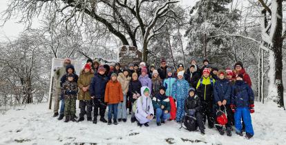 Na zdjęciu stoigrupa dzieci i dorosłych w zimowych kurtkach i czapkach. Do okoła śnieg, nad nimi osniezone gałęzie drzew, za nimi pomnik bohaterów powstania styczniowego(kamień z wyrytym napisem). 