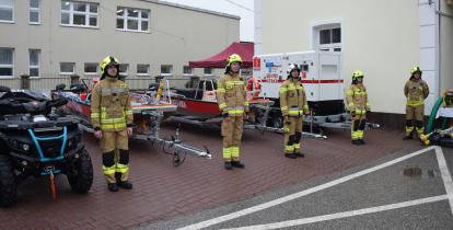 Na zdjęciu mężczyźni w mundurach strażackich stoja przed quadem, łodzią i przyczepą samochodową.