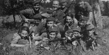 Czarno-białe zdjęcie. Na nim dziesięciu młodych męzczyzn w polskich mundurach z okresu II wojny światowej. W rękach trzymają karabiny, pozują do zdjęcia. Po prawej ich stronie leży na trawie młoda dziwewczyna, uśmiecha sie i tez pozuje do zdjęcia.