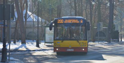 Ulica, po której jedzie czerwono-pomarańczowy autobus komunikacji miejskiej. Nad jego przednią szybą wyświetla się napis: 200. 