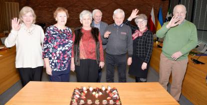 7 seniorów stoi obok siebie w rzędzie. Przed nimi na stole leży duży tort. 