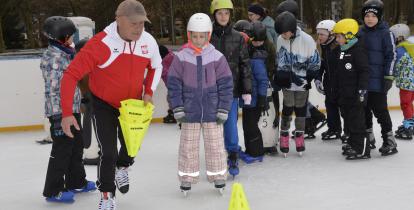 Na zdjęciu na lodowisku:  w pierwszym planie mężczyzna na łyżwach w biało-czerwonej kurtce; za nim stoją dzieci też na łyżwach, na lodzie, w kaskach. jedno za drugim.