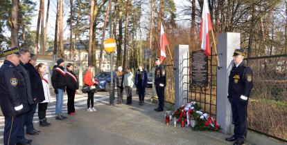 Grupa kilkunastu osób stoi przed pomnikiem pamięci, umieszczonym na ogrodzeniu Białego Dworku. Składają kwiaty i znicze. 