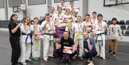 Młode dziewczyny i chłopcy w strojack zawodników karate. Na szyjach mają zawieszone medale, a w dłoniach dyplomy. Przed nimi kuca męzczyzna z pucharem w dłoni i kobieta.