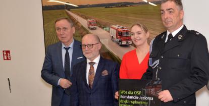 Na zdjęciu od prawej mężczyzna w strażackim mundurze i tabliczką w dłoniach z napisem napisem „Wóz dla OSP Konstancin-Jeziorna dzięki rządowemu wsparciu”. Obok kobieta w czerwonej sukience i dwóch mężczyzn w garniturach.