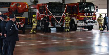 W hali stoją mężczyźni i kobiety w galowych mundurach strażackich, za nimi wozy ratowniczo-gaśnicze