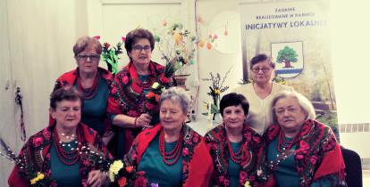 Siedem starszych kobiet w strojach ludowych, mają palmy wielkanocne w dłoniach. Za nimi baner z napisem: „Zadanie realizowane w ramach inicjatywy lokalnej”. 