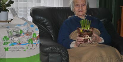 Na fotelu siedzi starsza kobieta. W dłoniach trzyma kwiaty.