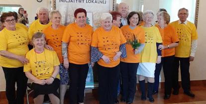 Grupa dwunastu starszych osówb, kobiet i męzczyzn. Sa w żółtych i pomarańczowych koszulkach. Za nimi jest plakat z napisem: zadanie realizowane w ramach inicjatywy lokalnej.