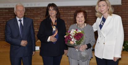 Na zdjęciu stoją trzy kobiety (jedna starsza trzyma w dłoniach bukiet kwiatów). PO lewej stronie stoi starszy mężczyzna, za nimi ceglana ściana i zawieszony ekranem.