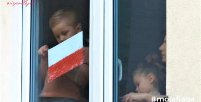 Dziecko stoi w oknie i trzyma w ręku biało-czerwoną flagę. 