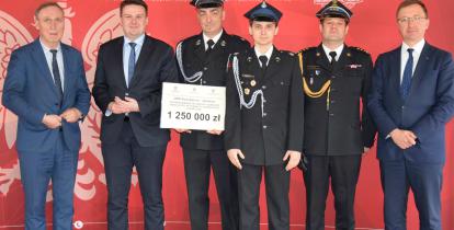 Sześciu mężczyzna, w tym trzech w mundurach strażackich, stoi na tle czerwonej ścianki promocyjnej Państwowej Straży Pożarnej. Jedna osoba trzyma prostokątną tabliczkę, na której widnieje kwota 1 250 000 zł.