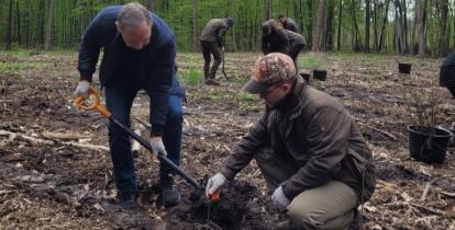 Dwóch mężczyzn, jeden pochylony z łopata, drugi kuca i trzyma sadzonkę drzewka. Za nimi inni ludzie oraz  drzewa.