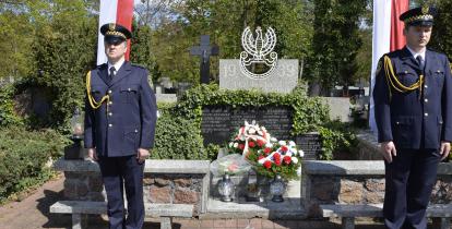 Dwóch mężczyzn w mundurach strazy miejskiej stoi czed grobem, który jest ogrodzeny niskim murkiem. Na tablicy wypisane są imiona i nazwiska oraz  napis „1939, poległym za ojczyznę”. 