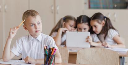 W sali za szkolnym stołem siedzi chłopiec w białej bluzce. Ma ołówek przy głowie. Na stole leżą zeszyty i kredki. Za nim, również za szkolnym stołem, siedzą trzy dziewczynki i patrzą w monitor komputera.