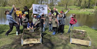 Dziei i dorośli nad brzegiem rzeki. Podnoszą do góry ręce. Z tyły dwóch mężczyzn trzyma plakat z napisem „NASZ BÓBR STOWARZYSZENIE”. Przed nimi donice z ziemia i gałązkami. Jedno dziecko leje na jedną z nich wodę z konewki.     