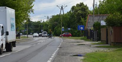 Zniszczona asfaltowa droga bez pobocza. Jedzie po niej ciężarówka, samochody osobowe oraz przechodzi przez nia mężczyzna z rowerem.