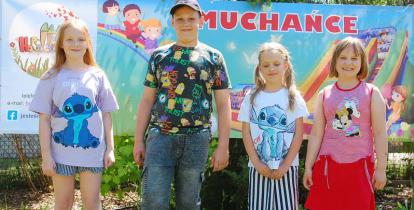 Postaci dzieci – trzy dziewczynki i jeden chłopiec stoją na trawie. Za nimi plakat z napisem: dmuchańce.