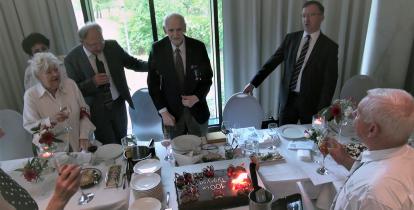 Za stołem stoi starszy mężczyzna, a obok niego stoją trzy osoby. Na stole stoi tort ze świeczkami.