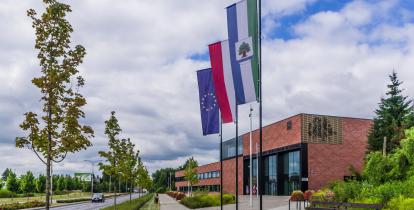 Duży, nowoczesny budynek użyteczności publicznej  ukazany z dużej perspektywy narożnika, ceglane ściany, duże okna i oszklenia, przed budynkiem drzewa.