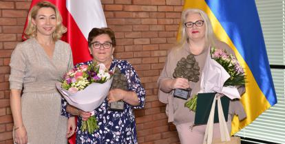 Trzy kobiety. Dwie z bukietami kwiatów w dłoniach. Za nimi biało-czerwona fla Polska oraz niebiesko-żółta Ukrainy oraz ceglana ściana i okno zasłonięte roletą.