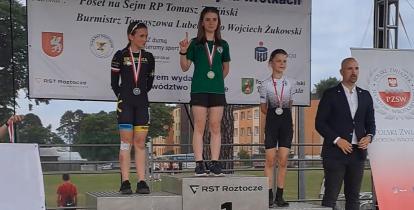 Podium. W środku stoi dziewczyna ze złotym medalem na szyi i zielonej bluzce. Po prawej i lewej jej stronie inne dziewczyny również z medalami na szyi (srebro i brąz).  Obok podium stoi młody mężczyzna w marynarce, dżinsach i białej koszuli.