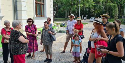 Przestrzeń publiczna. Grupa osób stoi przed budynkiem, patrzy na przewodnika.