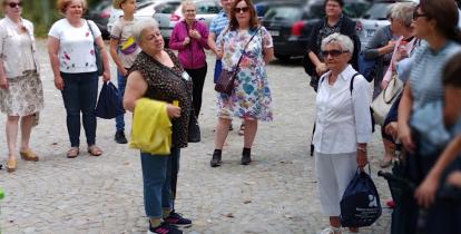 Przestrzeń publiczna. Grupa osób stoi przed budynkiem, patrzy na przewodnika.