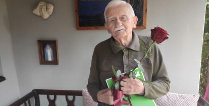 Staruszek trzyma w ręku dużą czerwoną róże i zieloną teczkę