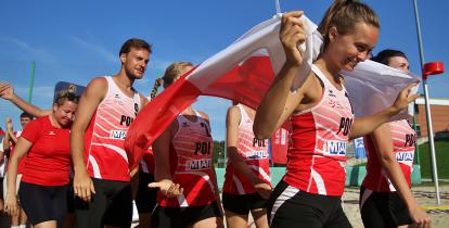 Młoda kobieta w stroju sportowym idzie na czele mężczyzn i kobiet, też w sportowych strojach, trzyma biało-czerwoną flagę.