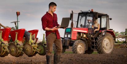Rolnik trzymający laptopa na tle maszyn rolniczych.